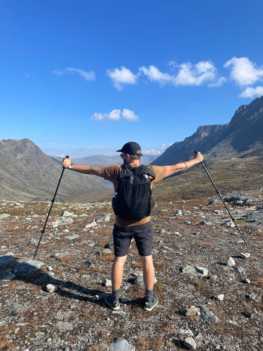 Man med utsträckta armar på en vandringsled med en ryggsäck och vy över fjäll